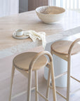Natural timber stools at a travertine breakfast bar styled with a sculptural ceramic bowl, handmade cup and plate, and soft textured cloth in a light-filled KULALA Living kitchen setting