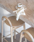 Minimalist kitchen styling featuring KULALA natural ashwood bar stools beneath a travertine benchtop, styled with a ceramic mug, plate and textured linen face washer