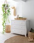 A serene nursery setting featuring woven storage baskets, a white wooden cot, a matching dresser, and a macrame hanging planter, creating a warm and natural space for a baby