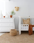 Serene nursery featuring a white dresser and cot, complemented by woven textures, a jute rug, rattan basket, natural decor accents, and a soft linen throw for a warm, organic aesthetic