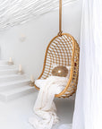 A natural rattan hanging chair with an open-weave design, suspended by a rope in a serene, all-white setting. Styled with plush white cushions, a textured throw, and a decorative sunburst pillow, creating a tranquil and inviting retreat