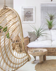 Natural rattan hanging chair with an intricate open-weave design, suspended in a bright boho-style lounge with a white cushion, rattan sofa, and wooden coffee table, creating a relaxed coastal aesthetic
