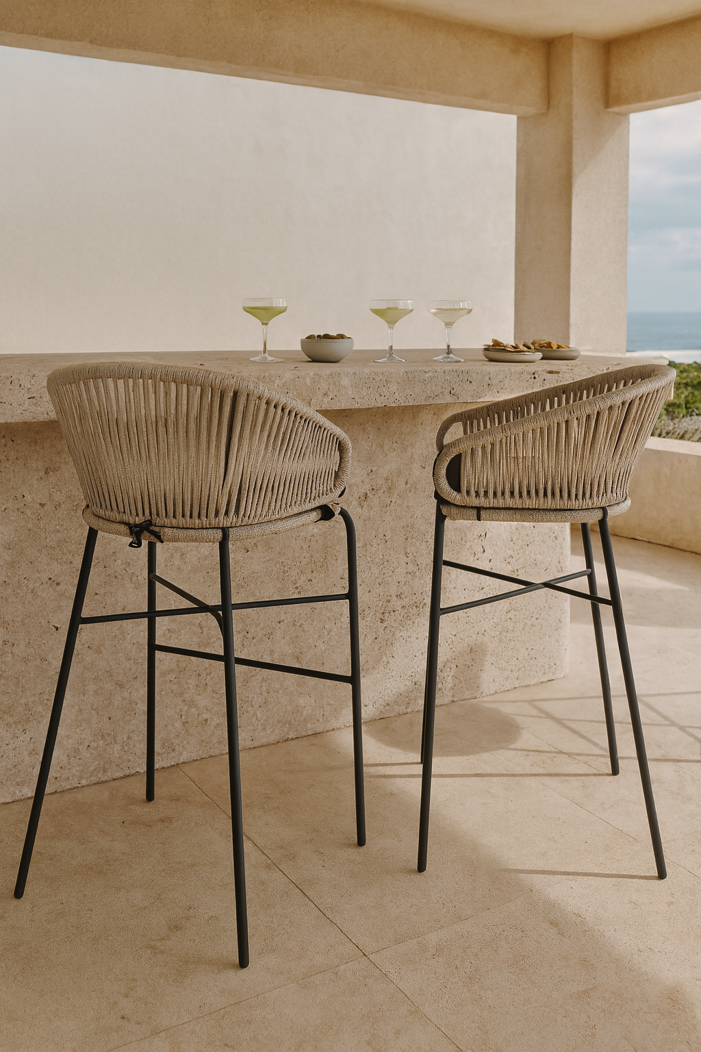 Natural rope outdoor bar stools with curved backs and black metal legs styled at a textured stone bar on a coastal balcony with margaritas and snacks