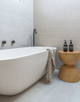 Modern bathroom featuring a freestanding stone bath, black tapware, and a carved magnesium oxide hourglass side table with Aesop bottles and a neutral-toned towel.