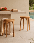 Pair of natural teak round stools styled by a stone bench in a poolside outdoor setting with neutral tones and relaxed summer decor