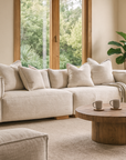 Neutral linen sectional sofa styled with scatter cushions, paired with a round oak coffee table in a light-filled living room overlooking the garden and pool