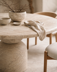 Nuru dining table in dune hydro dipped finish with round top and sculptural pedestal base styled in a soft neutral dining setting with natural textures.