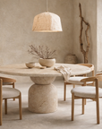 Nuru dining table in dune hydro dipped finish with round top and sculptural pedestal base styled in a soft neutral dining setting with natural textures.