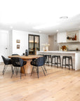 A stunning open-plan kitchen and dining area featuring a natural wood dining table surrounded by black woven chairs. The modern kitchen boasts a white island with matching black woven bar stools, gold fixtures, and a stylish tiled backsplash. The space is enhanced with warm timber flooring and curated decorative accents, creating a contemporary yet inviting atmosphere