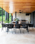 A stylish outdoor dining space featuring a black gable house with timber ceiling details, an open-plan living area, and a built-in wood-fired pizza oven. Surrounded by lush greenery, this modern retreat blends contemporary architecture with natural elements