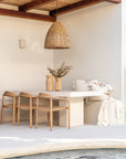 Outdoor dining setting featuring a teak chairs and travertine-look dining table beside a pool under a woven pendant light