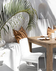 Outdoor dining setting with a white wicker chair and natural wood table, styled with earthy cushions and tropical foliage for a relaxed, coastal vibe.
