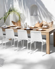 A serene outdoor dining setup by Kulala Living featuring a solid wood dining table paired with white wicker chairs, styled with earthy-toned cushions, ceramic vases, and natural tableware under soft dappled sunlight