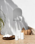 Serene outdoor dining space featuring a recessed white cement vase, wooden tableware, and soft neutral-toned cushions, bathed in natural sunlight and palm leaf shadows