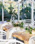 A beautifully styled outdoor dining space featuring a natural live-edge wooden table, white woven chairs, and elegant table settings with greenery and glassware, creating a refined yet relaxed entertaining area