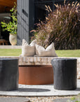 A modern outdoor setting featuring a rusted fire pit surrounded by black timber stools and textured linen cushions on a rustic brick ledge. The natural tones and organic textures blend seamlessly with the lush greenery, creating a serene and inviting space for outdoor relaxation