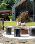A serene outdoor setting featuring KULALA Living’s black wooden stools arranged around a rusted steel firepit. Set against a modern black-clad home with lush greenery, this space blends contemporary design with natural elements for a stylish retreat. Neutral linen cushions add comfort to the inviting ambiance