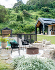 A tranquil outdoor firepit setting featuring modern black woven chairs, a rustic brick bench with linen cushions, and a lush hillside backdrop. This serene poolside space complements the contemporary black-clad home and open outdoor kitchen, creating a stylish and inviting retreat