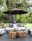 A beautifully styled outdoor lounge setting by Kulala Living, featuring teak armchairs and a sofa with white cushions, a rustic wooden coffee table, and black-and-white accent pillows under a large tree