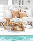 A stylish outdoor setting featuring a glass-top coffee table and side table with sculptural wooden bases, complemented by white lounge seating and woven cushions by the poolside.