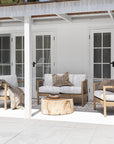 Modern coastal outdoor lounge setting with wooden accent chairs featuring white cushions and sleek spindle detailing, complemented by a round natural wood coffee table and dried floral arrangement