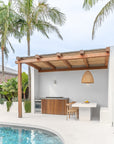 Modern outdoor poolside kitchen with natural wooden accents, woven pendant light, and minimalist concrete dining area surrounded by tropical palm trees.
