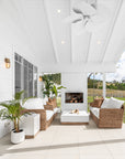 A serene outdoor patio featuring a stylish rattan sofa set with plush white cushions, a modern white coffee table with a wooden base, and a white brick fireplace. Surrounded by lush greenery, this inviting space blends coastal elegance with natural textures, perfect for relaxed outdoor living