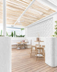 Outdoor dining area with natural wooden tables and woven seat chairs, shaded by a rustic wooden slat roof, surrounded by lush cactus plants and white walls.