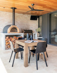 Modern outdoor dining setting with a square herringbone timber table, black chairs, woodfire pizza oven and BBQ, styled under a timber ceiling with ceiling fan.