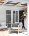 Modern coastal outdoor lounge setting with wooden accent chairs featuring white cushions and sleek spindle detailing, complemented by a round natural wood coffee table and dried floral arrangement