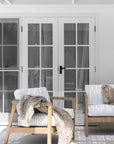 Modern coastal outdoor lounge setting with wooden accent chairs featuring white cushions and sleek spindle detailing, complemented by a round natural wood coffee table and dried floral arrangement