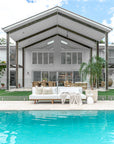 A stylish outdoor white sofa with plush cushions, set by a serene poolside with a modern home backdrop. The inviting space is accented with neutral decor, textured throws, and natural wood elements, perfect for relaxed outdoor living