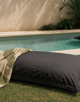 Playa Outdoor Bean Bag pillow in charcoal styled poolside with striped towel and matching floating bean bag in water.