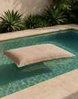 Playa Outdoor Bean Bag pillow in sand colour floating in a turquoise pool with surrounding tropical greenery.