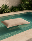 Playa Outdoor Bean Bag pillow in sand colour floating in a turquoise pool with surrounding tropical greenery.