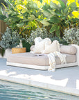 Poolside daybed with neutral cushions and throw, paired with a wooden side table, surrounded by tropical greenery for a tranquil outdoor lounging space.