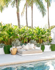 Tranquil poolside scene featuring a low-profile outdoor daybed with neutral cushions, lush tropical greenery, and a magnesium oxide stool styled with glassware and a throw.