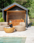 Poolside lounge area featuring two contemporary armless outdoor chairs in textured neutral fabric and a solid round magnesium oxide coffee table, set in front of a timber-lined sauna with glass doors.
