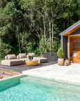 Poolside scene featuring two upholstered outdoor daybeds and a round magnesium oxide coffee table on a raised deck beside a sauna, surrounded by lush green forest
