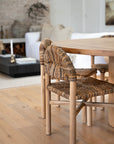A close-up of a rattan and timber dining chair with an intricate handwoven seat and backrest, styled with a solid wood dining table in a neutral-toned living space. The warm, earthy textures enhance the organic modern aesthetic of the room