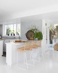 A modern coastal kitchen featuring three handwoven rattan bar stools with white metal frames, styled against a white island bench. The space is accented with tropical greenery, textured decor, and natural elements, creating an inviting and relaxed aesthetic