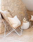 A pair of rattan sling chairs with white metal frames styled in a modern coastal interior, set against a natural stone wall with soft neutral décor for a relaxed and inviting aesthetic