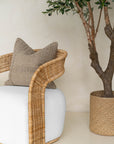 A close-up of a rattan armchair with a white upholstered seat, styled with a textured linen cushion. Paired with a woven basket planter and an olive tree, this setting exudes natural elegance and relaxed sophistication