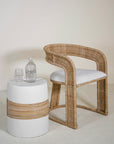 A natural rattan dining chair with a curved backrest and white cushion, styled alongside a white and rattan-accented side table with glassware. The minimalist setting highlights organic textures, perfect for modern coastal or relaxed interiors