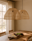 Pair of woven rattan pendant lights hanging above a wooden dining table with a cheese and fruit platter, styled in a sunlit dining room.