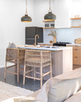 Pair of natural rattan bar chairs styled at a timber kitchen island with brass tapware, ceramic pendants, and coastal-inspired decor for a relaxed contemporary loo