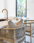 Natural rattan counter stools at a white stone breakfast bar with neutral cushions and warm brass tapware in a modern kitchen setting