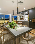 Contemporary dining room with a concrete dining table and wooden chairs featuring woven seats, overlooking a city skyline at night through large glass doors.