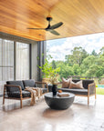 Outdoor setting featuring two timber-framed rope sofas with black cushions, styled with neutral throws and pillows, complemented by a round black coffee table on a light stone patio overlooking lush greenery.