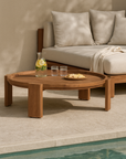Round teak outdoor coffee table styled with a jug of lemon-infused water and glasses, positioned beside a cushioned daybed on a poolside terrace.